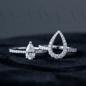 May include: A silver-toned ring set featuring a pear-shaped center stone on a pavé band alongside a matching open-frame pear-shaped contour band encrusted with small clear stones. The jewelry is displayed against a dark, textured surface.