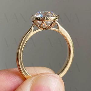 May include: A gold engagement ring with a large, clear, round-cut diamond. The ring features a delicate, openwork design around the diamond and a smooth, polished band. The ring is held by a person's fingers.
