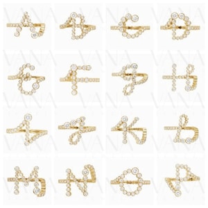 May include: A collection of gold-toned rings, each designed as a different letter of the alphabet. The letters are formed with small, round, clear gemstones. The rings are displayed on a white surface.