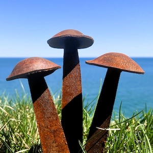 May include: Three rusty metal mushrooms are arranged in a triangular formation in a bed of green grass. The mushrooms are tall and slender, with rounded tops. The background is a blue sky and ocean.