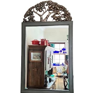 May include: A rectangular mirror with a dark gray frame and a decorative bronze-colored tree design at the top. The mirror reflects a kitchen scene with red pots and blue glass objects.