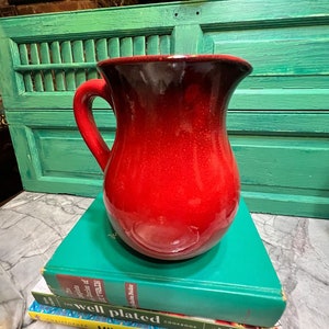May include: A red ceramic pitcher with a black rim and a handle. The pitcher is sitting on top of a stack of three cookbooks. The top book is green and says "The Well Plated Cookbook". The middle book is yellow and says "Milk Street Cookbook". The bottom book is blue and says "Tuesday Night  Hannah  Recipes".