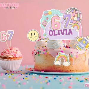 Puede incluir: Un pastel y cupcakes decorados con glaseado rosa y chispas. El pastel tiene un adorno que dice "I was 67 Now I'm Olivia" con una carita sonriente, un arcoíris y un cohete. Los cupcakes tienen un adorno "67".