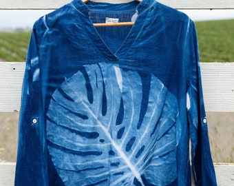 May include: A blue long-sleeved shirt with a white leaf design printed on the front. The shirt has a v-neck and is hanging on a wooden hanger.
