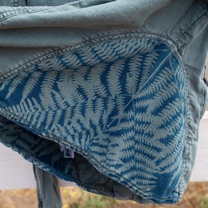 Puede incluir: Una chaqueta vaquera azul con un patr&oacute;n de helechos en el forro interior. La chaqueta est&aacute; doblada y colocada sobre una valla de madera blanca.