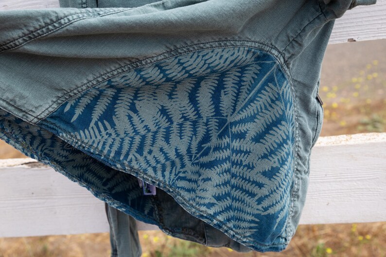 Puede incluir: Una chaqueta vaquera azul con un patr&oacute;n de helechos en el forro interior. La chaqueta est&aacute; doblada y colocada sobre una valla de madera blanca.
