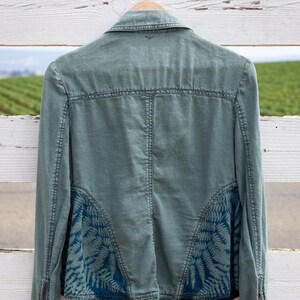 Puede incluir: Una chaqueta vaquera verde con un estampado de helechos azules en la espalda. La chaqueta tiene cierre de cremallera y mangas largas.