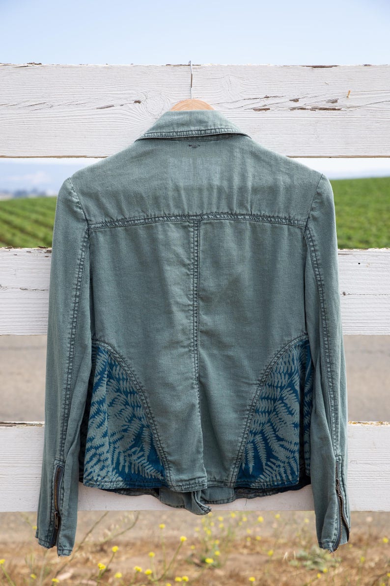 Puede incluir: Una chaqueta vaquera verde con un estampado de helechos azules en la espalda. La chaqueta tiene cierre de cremallera y mangas largas.