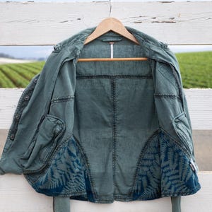 Puede incluir: Una chaqueta vaquera azul verdosa con un forro de estampado de helechos. La chaqueta est&aacute; colgada en una valla de madera blanca.