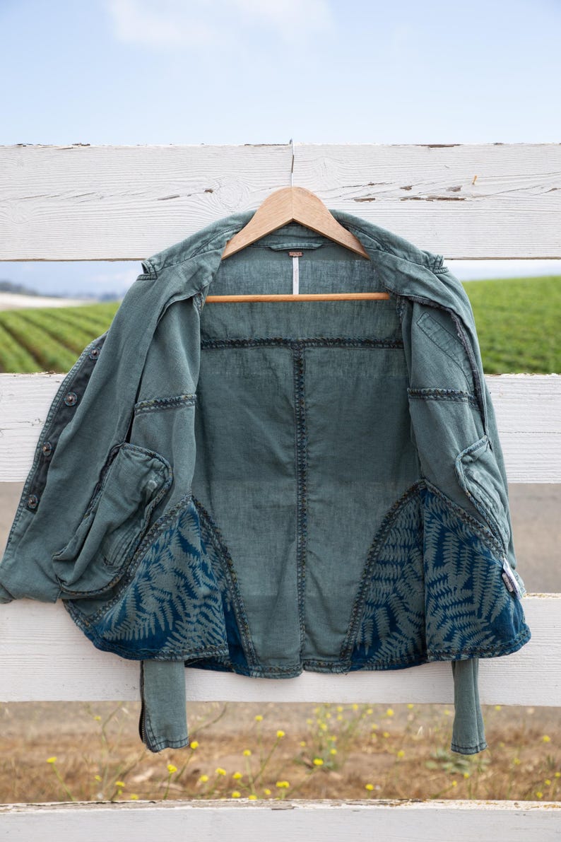 Puede incluir: Una chaqueta vaquera azul verdosa con un forro de estampado de helechos. La chaqueta est&aacute; colgada en una valla de madera blanca.