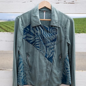 Puede incluir: Una chaqueta vaquera verde con un estampado de helechos azules en la parte delantera y las mangas. La chaqueta tiene cierre de cremallera y cuello.