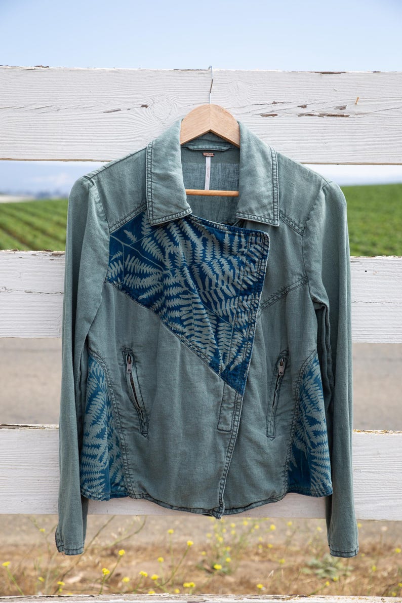 Puede incluir: Una chaqueta vaquera verde con un estampado de helechos azules en la parte delantera y las mangas. La chaqueta tiene cierre de cremallera y cuello.