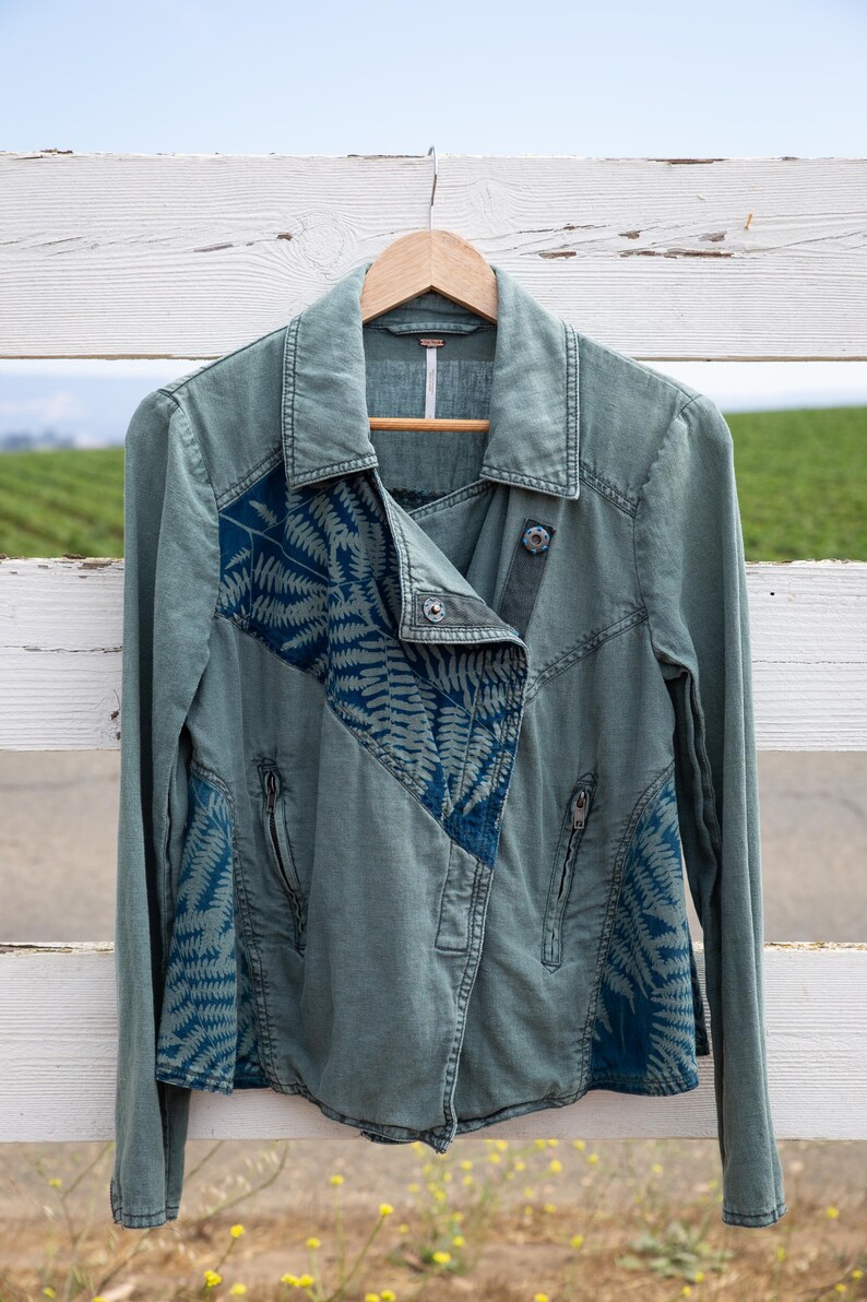 Puede incluir: Una chaqueta vaquera verde con un estampado de helechos azules en las mangas y el frente. La chaqueta tiene cierre de cremallera y cuello.