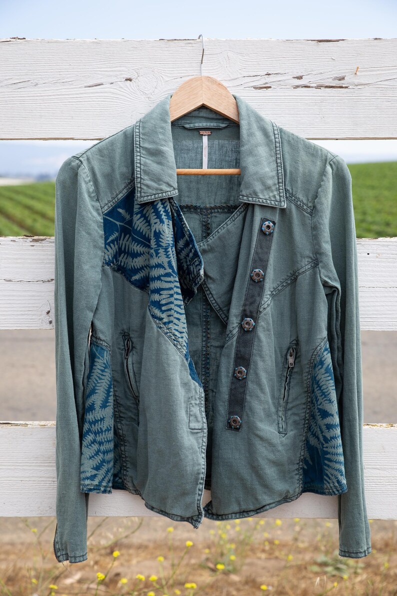 Puede incluir: Una chaqueta utilitaria verde con un estampado floral azul en las mangas y la espalda. La chaqueta tiene una cremallera y un cierre de botones en la parte delantera.