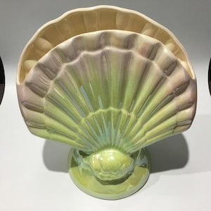 May include: A ceramic seashell-shaped ornament in shades of green, lavender, and cream. The fan-shaped design sits on a round base. The piece has a glossy finish and is likely a decorative item for a coastal or nautical-themed home.