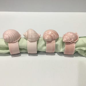 May include: A set of four pale pink ceramic napkin rings, each featuring a distinct seashell design on top. The rings are arranged in a horizontal row, holding a light green fabric napkin that is folded through the centre of each ring.