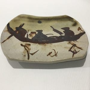 May include: A rectangular ceramic plate with a light beige, speckled surface. The centre features a dark brown, hand-painted illustration of a boat with figures, accented by abstract calligraphic brush strokes in the same dark brown tone.