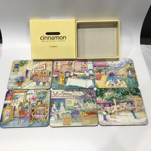 May include: A set of six square coasters featuring watercolour illustrations of European street scenes, including cafes and shops. The coasters are displayed with a yellow box that reads "cinnamon" and "Making Art Your Lifestyle."
