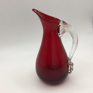 May include: A deep red glass jug with a clear glass handle. The jug has a rounded body and a flared spout. The handle is attached to the body with decorative glass elements. It is likely used for serving drinks.