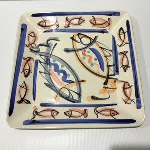 May include: A square ceramic plate with an off-white base featuring hand-painted fish motifs in peach, blue, and black. The border is decorated with a repeating pattern of small fish, while the centre displays two larger stylised fish designs.