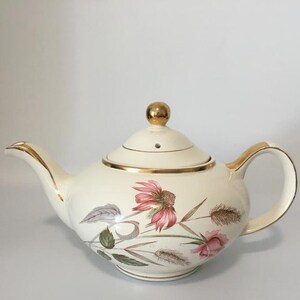 May include: Cream-coloured ceramic teapot with a gold handle, spout, and trim. The teapot features a floral design with pink and brown flowers, green leaves, and grey accents. The lid has a gold knob.