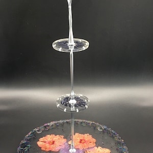May include: A clear, three-tiered serving stand with a decorative handle. The bottom tier features orange floral designs and a leaf, with iridescent beads around the edge. The middle and top tiers are clear with dark blue accents. Perfect for displaying cakes or pastries.