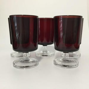 May include: Six burgundy-coloured stemmed glasses. The glasses have a cylindrical shape with a clear, circular base. The glass colour is a deep, rich red, and the stems are clear.