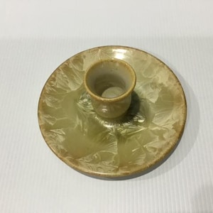 May include: A ceramic candle holder featuring a circular base with a light green and cream crystalline glaze pattern. A small, cylindrical cup is centred on the plate, matching the same glossy, variegated finish as the base.