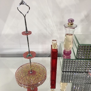 May include: A multi-tiered serving stand with pink glitter and gold details, topped with a decorative handle. A red perfume bottle and a clear perfume bottle with a purple top are also visible. The items are displayed on a mirrored surface.