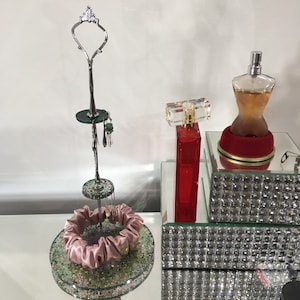 May include: A silver jewellery stand with a decorative top, holding earrings and a pink scrunchie. A red perfume bottle with a clear, rectangular top and a perfume bottle in the shape of a woman's torso are also visible.