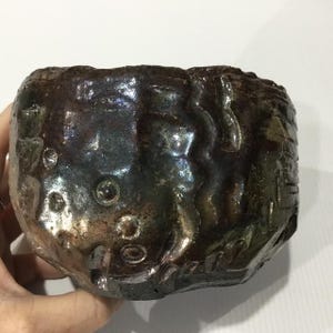 May include: A dark, metallic-toned ceramic bowl with a highly textured, uneven surface. The exterior features raised, organic ridges, circular indentations, and a glossy, iridescent glaze that shifts between deep brown, bronze, and charcoal hues.