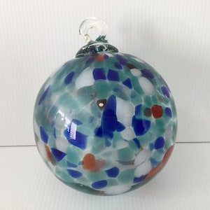 May include: A spherical glass ornament featuring a speckled pattern of teal, royal blue, white, and burnt orange flecks throughout the clear glass body. The ornament includes a small, integrated clear glass loop at the top for hanging purposes.