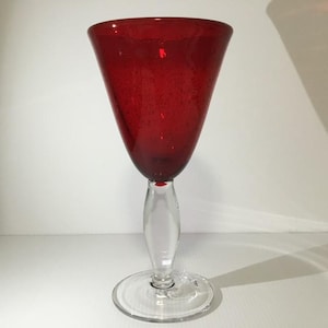 May include: A goblet with a deep red glass bowl featuring subtle internal air bubbles. The stem is made of clear glass with a rounded, bulbous centre section, and it rests on a wide, flat, circular clear glass base for stability.