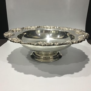May include: A silver-coloured decorative bowl with a wide, ornate rim featuring a floral design. The bowl has a hammered texture and sits on a pedestal base. The interior is highly reflective.