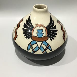 May include: A ceramic vase with a cream-coloured body, a black base, and a black rim. The vase features a hand-painted design of a winged figure with blue and brown accents. The vase is approximately 20 cm tall.