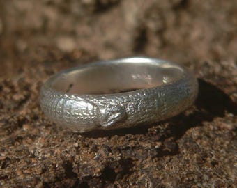 May include: A silver-coloured ring featuring a textured surface that mimics the appearance of snake scales. The band has a rounded profile and includes a small, raised detail resembling a snake head integrated into the continuous scale pattern.