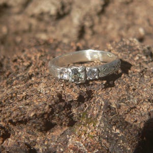 May include: A silver ring featuring a three-stone setting with a central round gemstone flanked by two smaller round stones. The metal band displays an intricate, engraved leaf-like pattern along its surface, providing a textured appearance.
