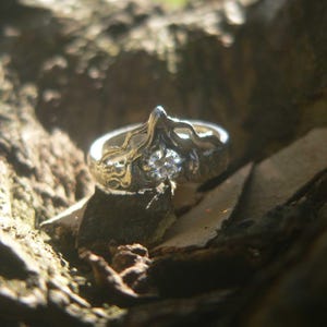 May include: A silver ring featuring a central round clear gemstone set within a band that has an organic, textured, and raised wave-like design. The metal surface appears polished in some areas and darkened in the recessed, detailed sections.