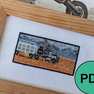 May include: A cross-stitch embroidery framed in a light brown wood frame. The embroidery depicts a gray truck with a trailer hauling a load of logs. The truck is driving on a dirt road with a blue sky in the background.