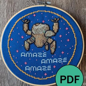 Könnte beinhalten: Ein Kreuzstich-Stickrahmen mit einer pixeligen Figur in Braun- und Beigetönen auf blauem Hintergrund. Das Wort "AMAZE" wird dreimal in Weiß wiederholt, mit rosa Sternakzenten. Der Rahmen befindet sich in einem Holzrahmen.