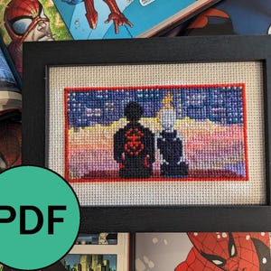 May include: Cross-stitch artwork in a black frame, depicting two figures against a sunset. One figure has a red and black design. The background includes comic books. The artwork is a unique piece of home decor.