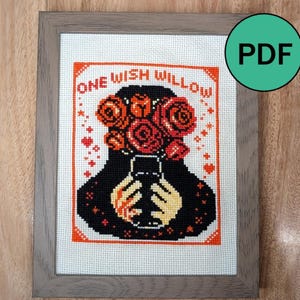 May include: A cross-stitch pattern in a gray frame featuring a black silhouette of a person holding a bouquet of red and orange roses. The text ONE WISH WILLOW is stitched in orange above the figure, surrounded by small orange hearts and stars.