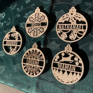 Peut inclure: Cinq boules de Noël en bois avec des motifs découpés et des noms : "JULES", "NATHANAEL", "BOUBOU", "LOUANE" et "MAMAN". Chaque boule a une forme unique, avec des flocons de neige, des feuilles et un renne. Les boules sont de couleur marron clair.