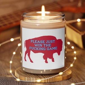 May include: A brown candle with a white label that says "PLEASE JUST WIN THE FUCKING GAME". The label also features a red silhouette of a buffalo. The candle is lit and sitting on a wooden coaster.