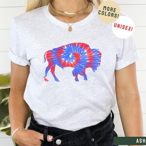 May include: A white t-shirt with a tie-dye buffalo silhouette in red and blue. The shirt is unisex and available in more colors. The word "ASH" is printed in green on the bottom right corner of the image.