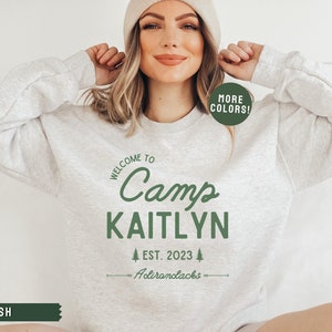 May include: A gray sweatshirt with the text "Welcome to Camp Kaitlyn Est. 2023 Adirondacks" in green. The sweatshirt has a green circle with the text "More Colors!" in white.