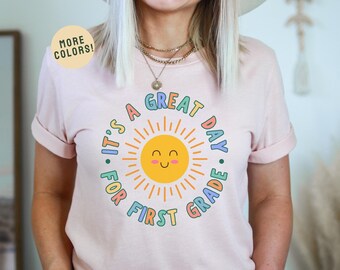 May include: Pink t-shirt with a colorful graphic that says "It's a Great Day for First Grade" with a smiling sun in the center.