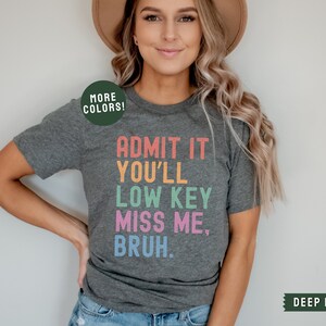 May include: A dark heather grey t-shirt with the text "ADMIT IT YOU'LL LOW KEY MISS ME, BRUH." printed in rainbow colors. The text is stacked vertically with each word in a different color.