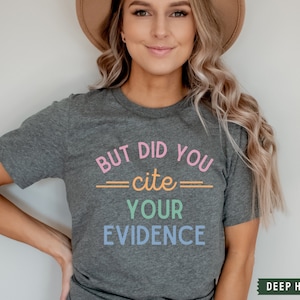 Op de afbeelding: Een grijs T-shirt met de tekst "BUT DID YOU cite YOUR EVIDENCE" in regenboogkleuren.
