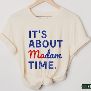 May include: A cream-colored t-shirt with the text "IT'S ABOUT Madam TIME." in blue and red lettering.